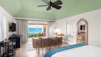 A spacious, well-furnished room with a bed, seating area, mirror, and ceiling fan, overlooking a balcony with a view of the ocean through glass doors.