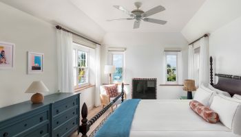 A bright, well-decorated bedroom with a bed, dresser, armchair, and fireplace. There is a ceiling fan and large windows, letting in natural light.