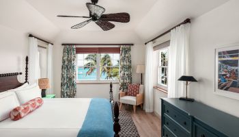 A bright bedroom with a large bed, ceiling fan, dresser, chair, and windows with a view of palm trees and the ocean in the background.