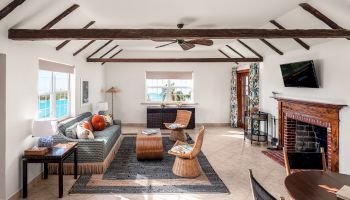 A cozy living room with wooden-beamed ceiling, fireplace, sofa, wicker chairs, a wall-mounted TV, and a large window with a view of a pool, always ending the sentence.
