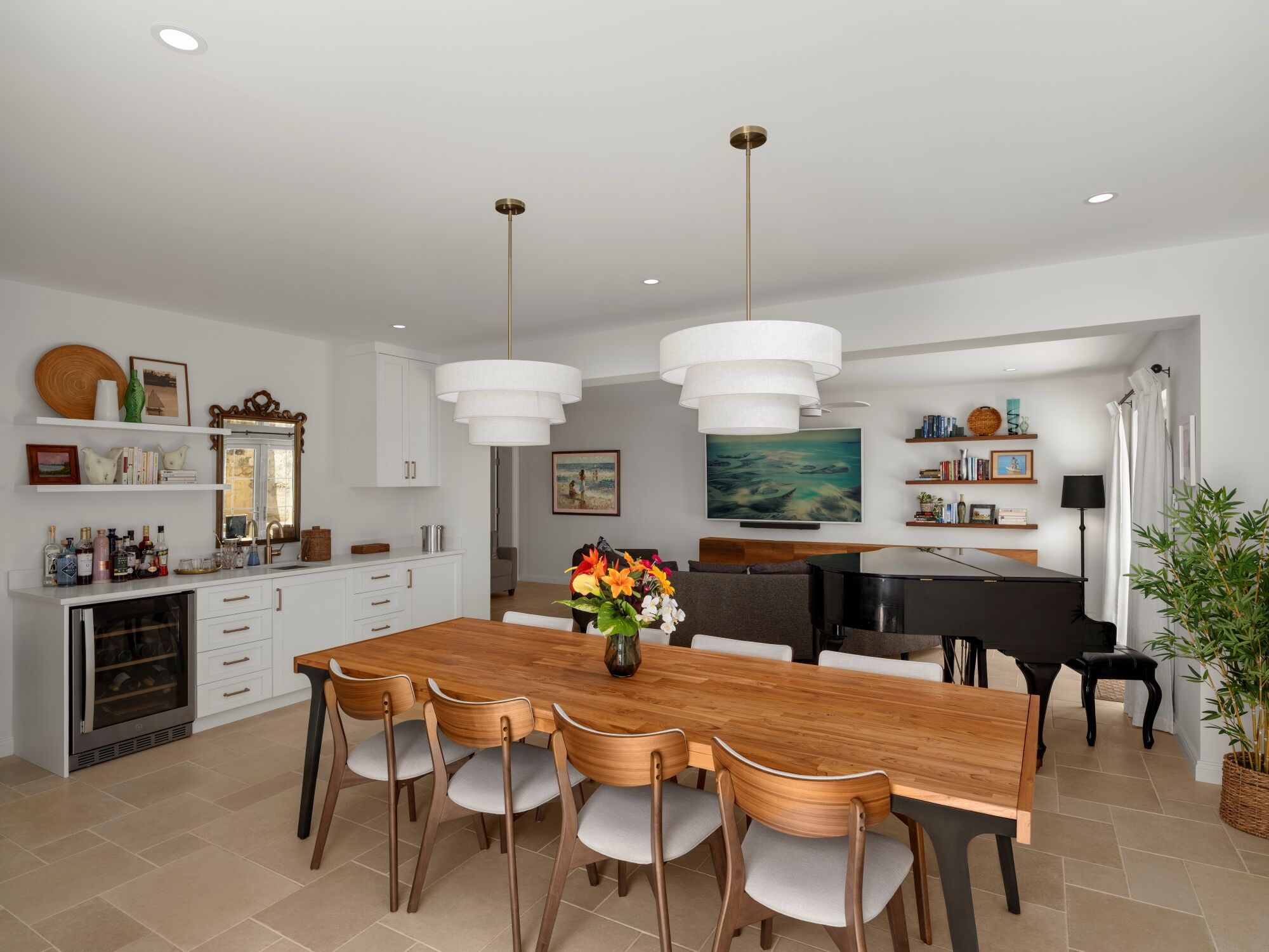 A bright, modern dining room with a long wooden table, six chairs, a built-in kitchen area on the left, and a living area with shelves and a piano on the right, plus plants and overhead pendant lights.