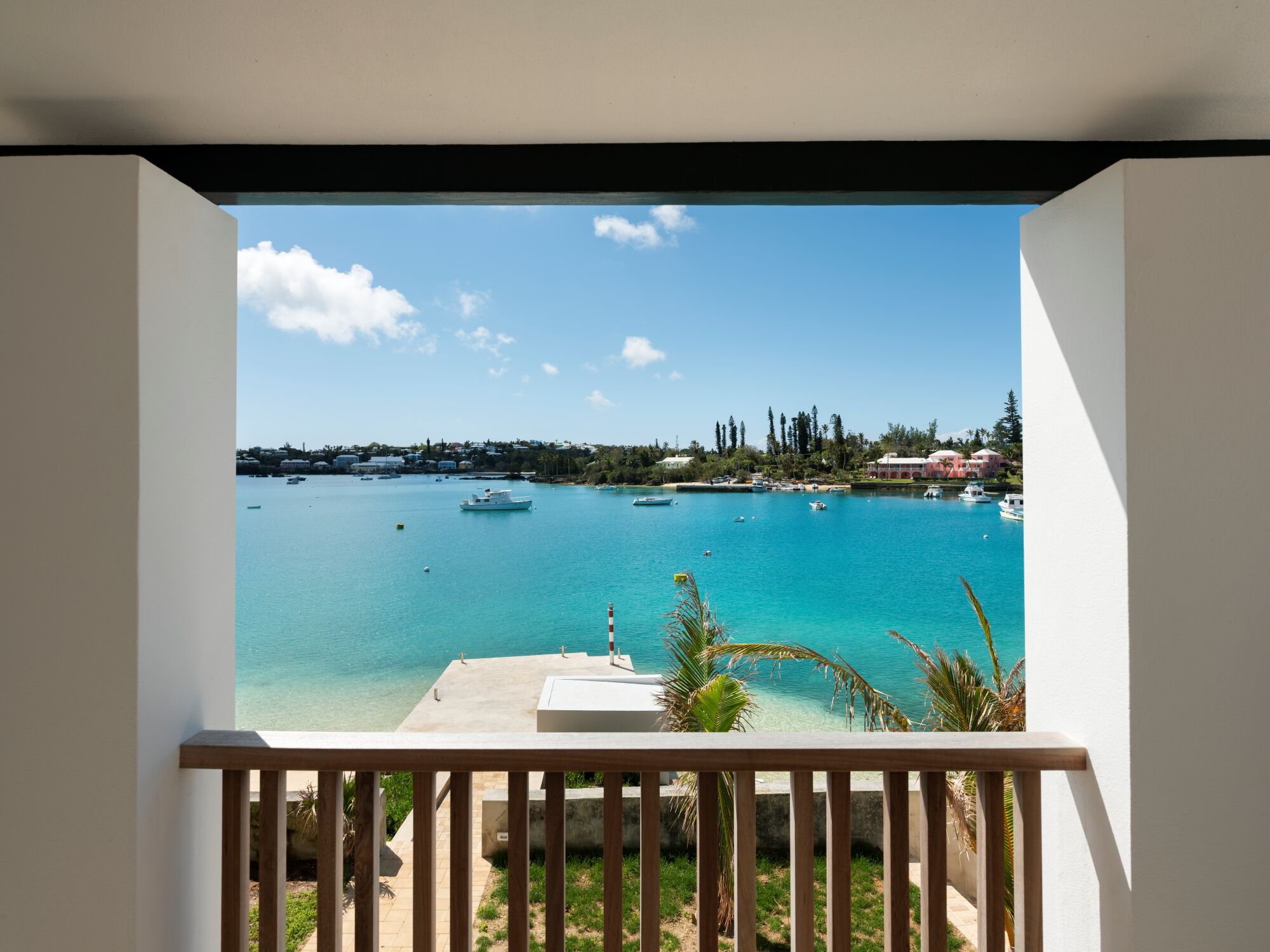 A balcony view opens to a turquoise bay with boats, white sand, palm trees, and a distant shore under a bright blue sky.