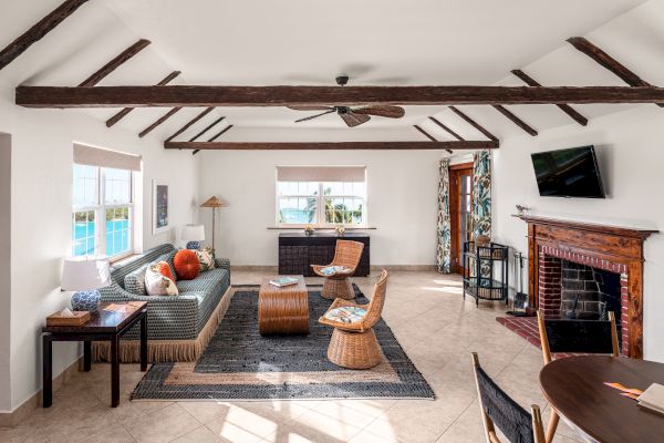 A cozy living room with exposed beams, a fireplace, a TV, and rattan furniture. There are multiple windows bringing in natural light.