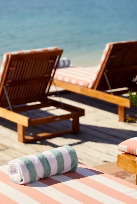 Sun loungers on a sunny beach deck with striped cushions, a small table of drinks, and rolled towels by the water.