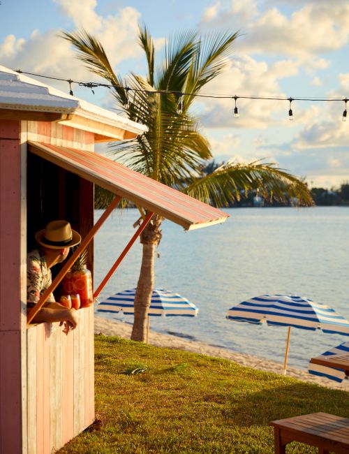 A person in a hat looks out from a small pink shack by the water, with palm trees and striped umbrellas in the background.