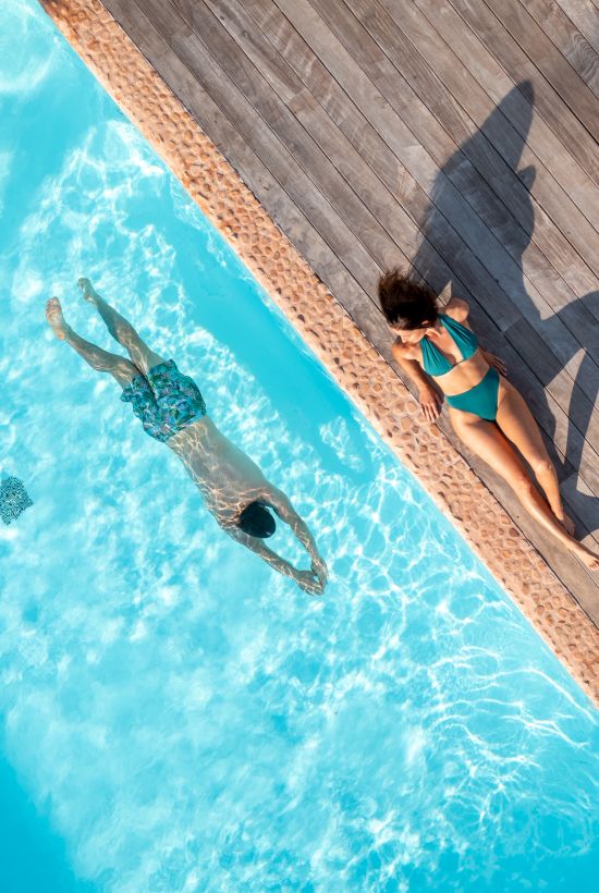 A person is swimming in a clear blue pool while another lounges on the deck, casting a shadow.