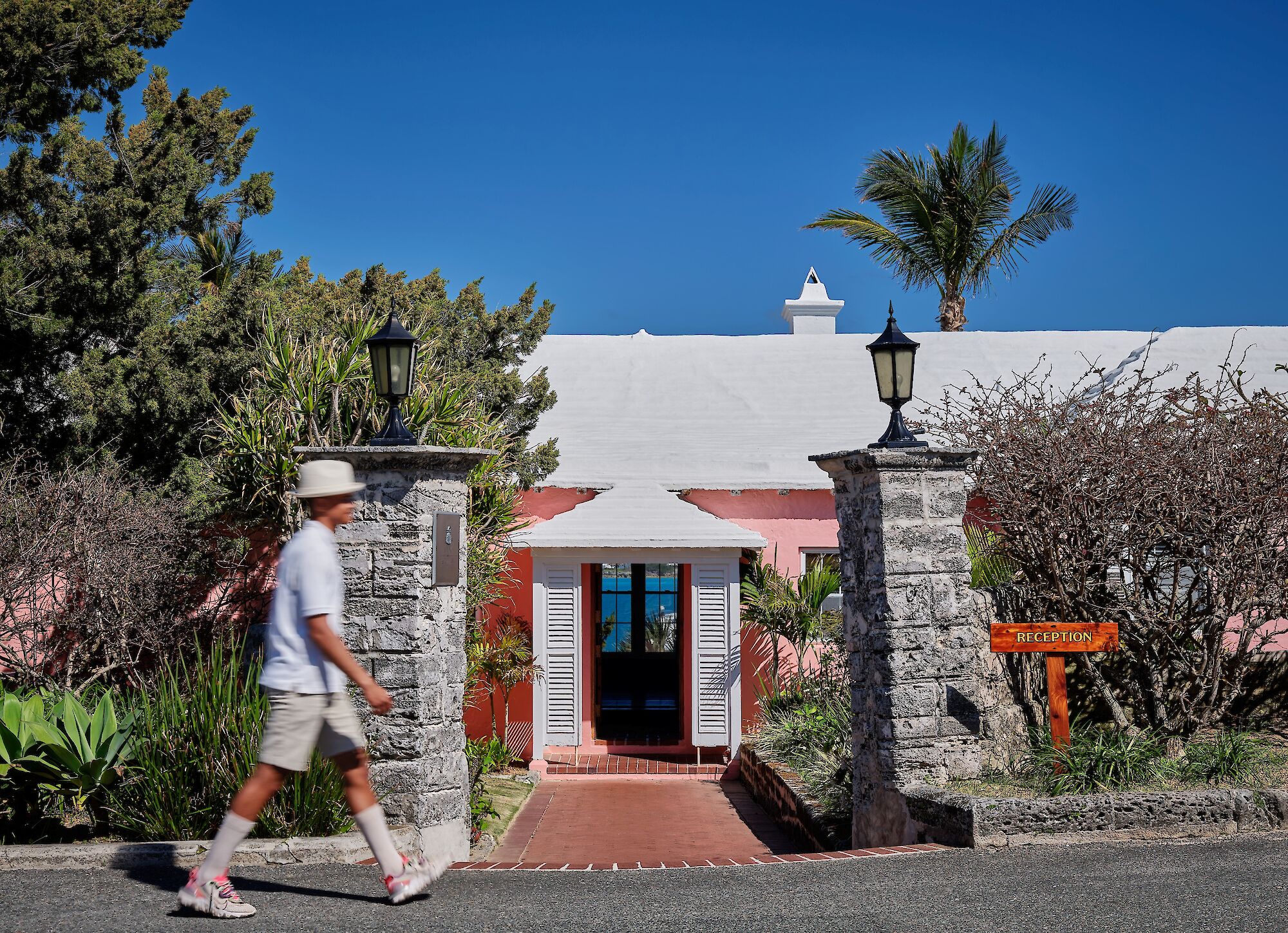 A tropical resort reception entrance with whitewashed walls, lantern posts, palm trees, and a guest in a hat walking by.