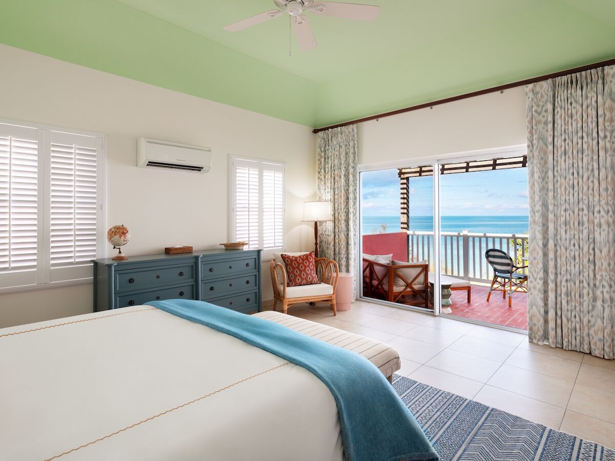 A bright bedroom with a ceiling fan, dresser, and shutters, plus a sliding door to an ocean-view balcony with patio chairs.
