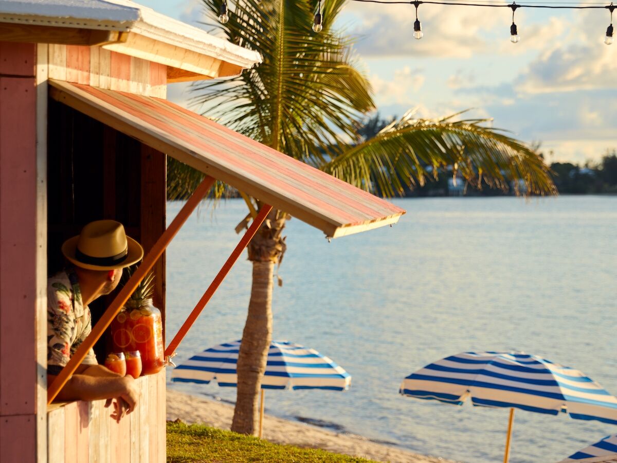 Beach hut deck with a hammock, palm tree, striped umbrellas, and string lights over a calm ocean under a sunset sky.