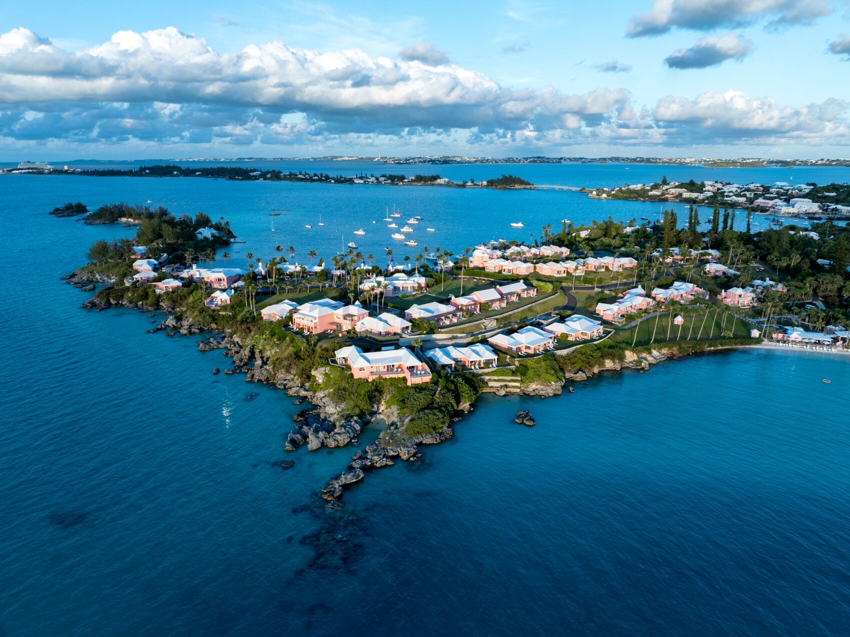 An aerial view of a tropical island resort with white-roofed villas, lush palm trees, and boats anchored around a tranquil blue lagoon.