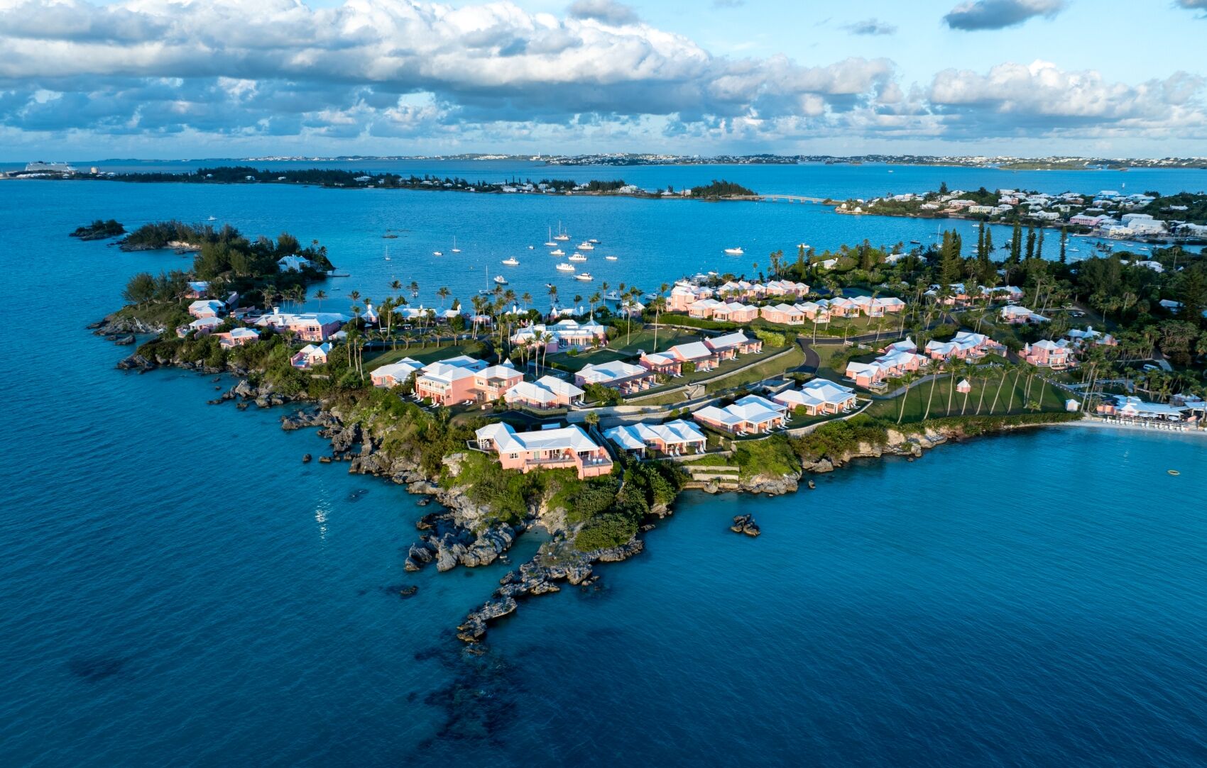 An aerial view of a tropical island resort with white-roofed villas, lush palm trees, and boats anchored around a tranquil blue lagoon.