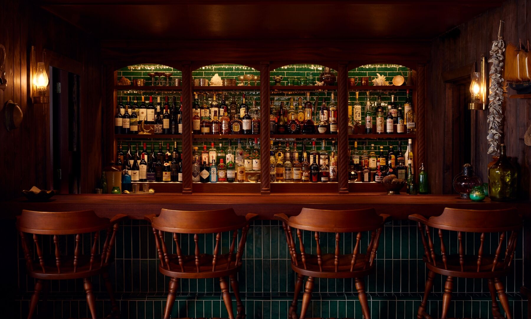 A cozy, dimly lit bar with a wooden counter, multiple bar stools, and shelves stocked with bottles behind the counter, evening vibe.