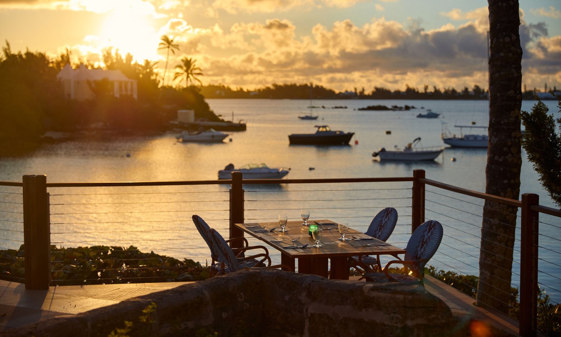Sunset over a tranquil harbor with boats, a wooden deck, and a table for two, palm trees, and warm golden light reflecting on the water.