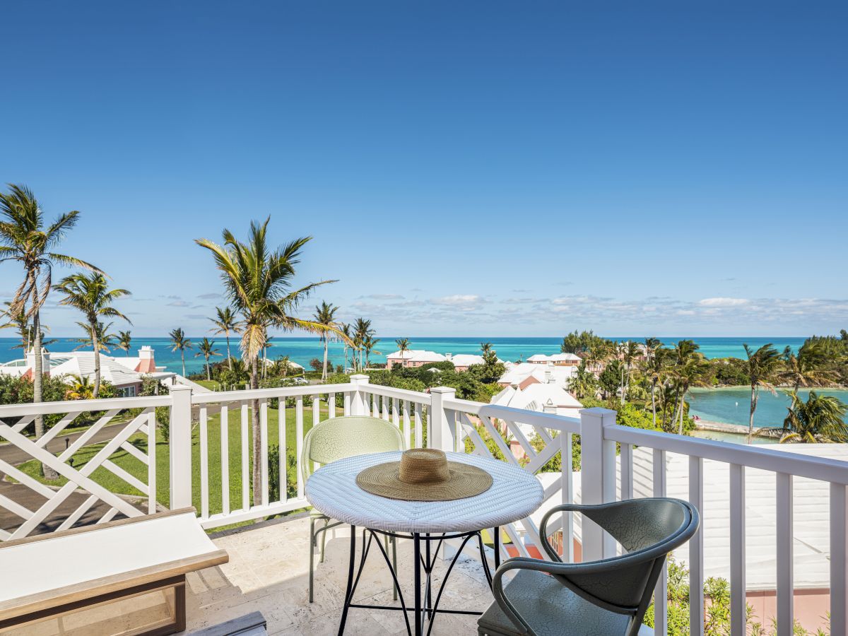 A sunny patio with a white table, a straw hat, two chairs, and a beautiful ocean view with palm trees and blue skies in the background.