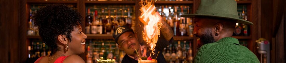 Two stylish people chat at a bar, a bartender with a flaming torch behind them; a warm, tropical night vibe with green-lit counter.