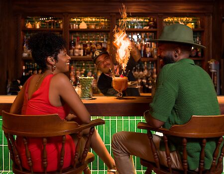 Two stylish people chat at a bar, a bartender with a flaming torch behind them; a warm, tropical night vibe with green-lit counter.