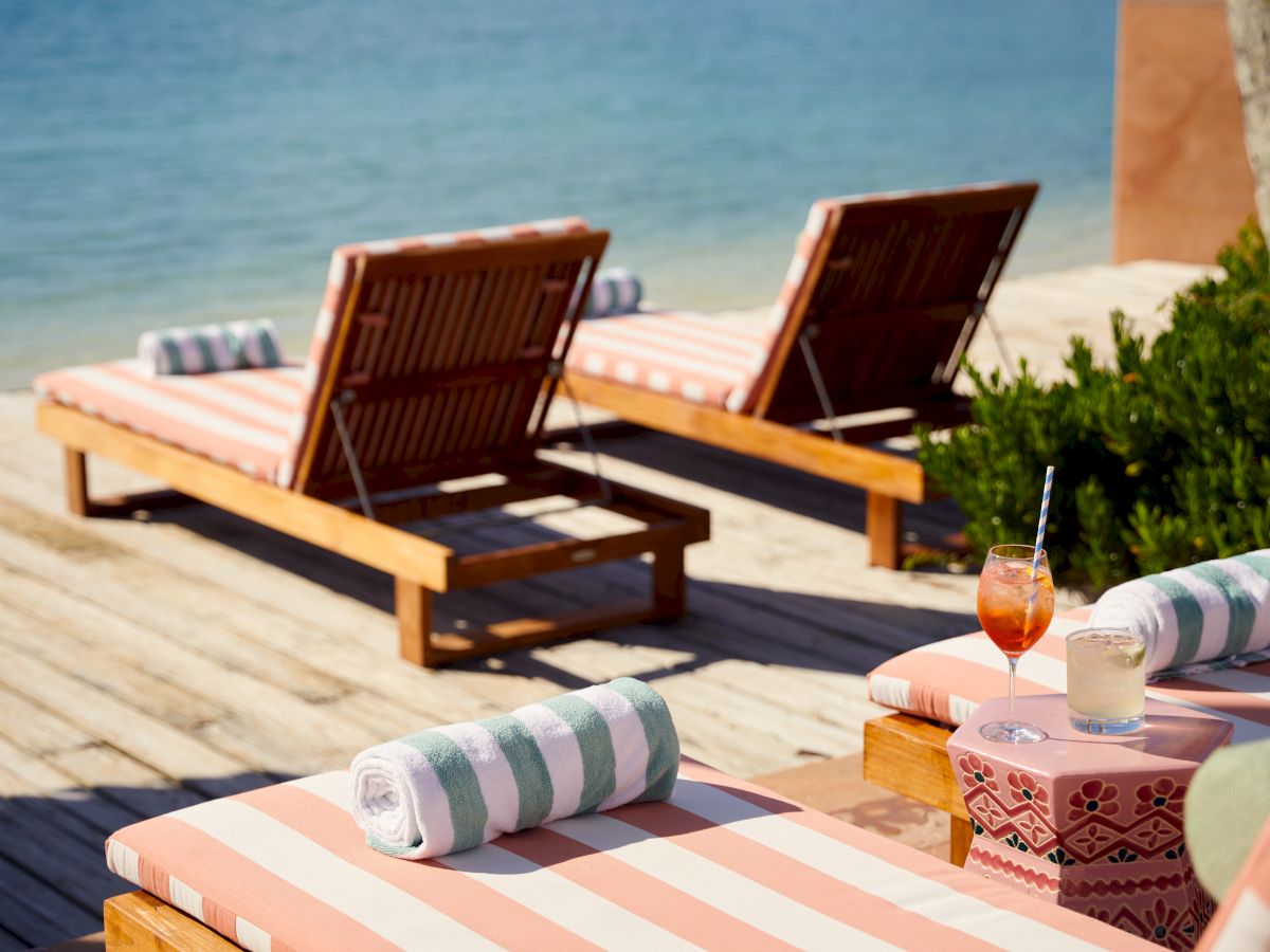 Two lounge chairs with striped cushions and rolled towels, positioned next to a beach with drinks on a small table nearby, by the water.