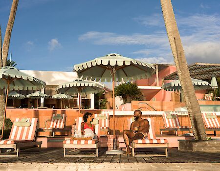 Two people lounge under striped umbrellas at a sunny poolside with pink walls, palm trees, and wicker chairs.