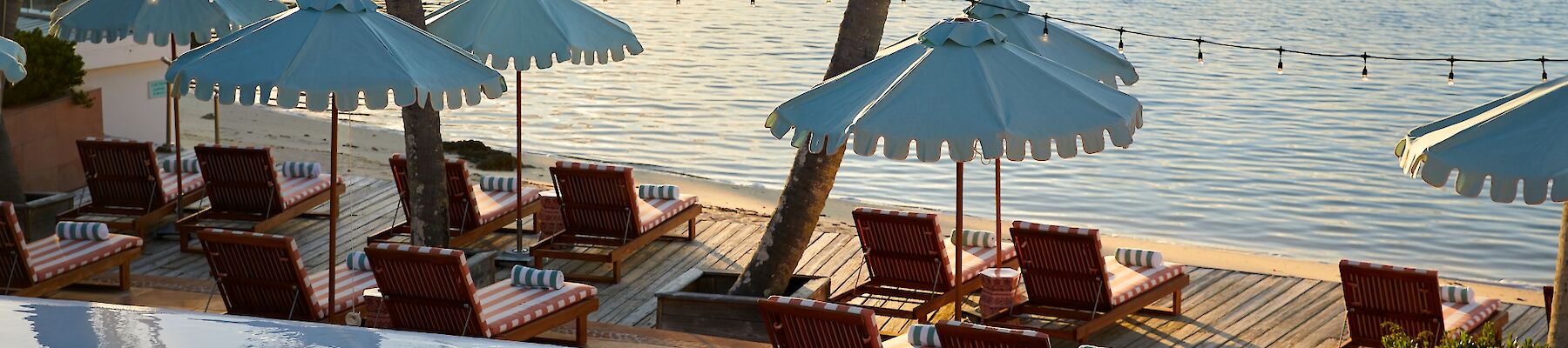 A tropical beach scene with blue umbrellas, sun loungers along a wooden deck, palm trees, and calm water with boats in the distance, at sunset.