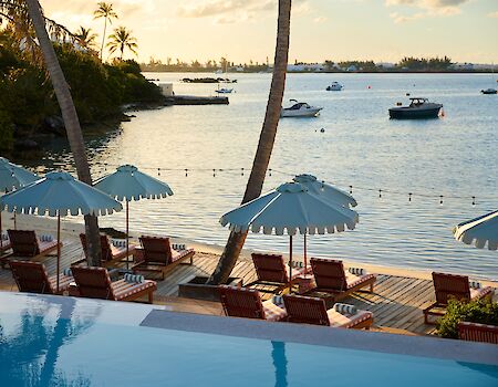 A tropical beach scene with blue umbrellas, sun loungers along a wooden deck, palm trees, and calm water with boats in the distance, at sunset.