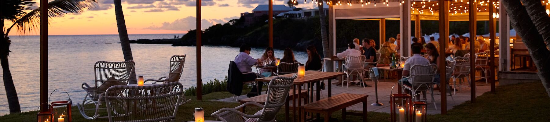 A seaside outdoor restaurant at sunset with string lights, palm trees, wicker chairs, lanterns, and diners by the water.