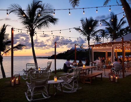 A seaside outdoor restaurant at sunset with string lights, palm trees, wicker chairs, lanterns, and diners by the water.