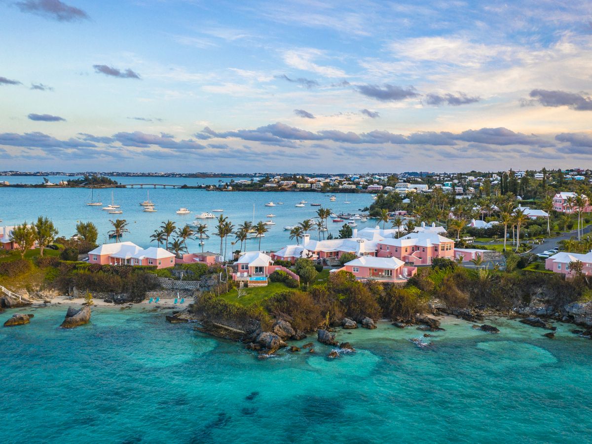 The image shows a coastal scene with turquoise water, pink houses, palm trees, and boats on the water, under a partly cloudy sky.