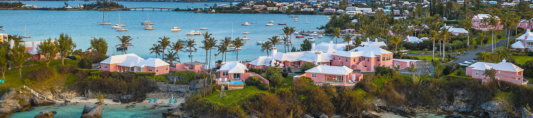 Colorful coastal scene with turquoise water, rocky shoreline, and a cluster of white and pastel houses along the coast at sunset.