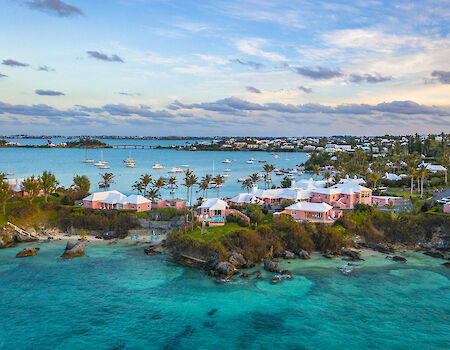 Colorful coastal scene with turquoise water, rocky shoreline, and a cluster of white and pastel houses along the coast at sunset.