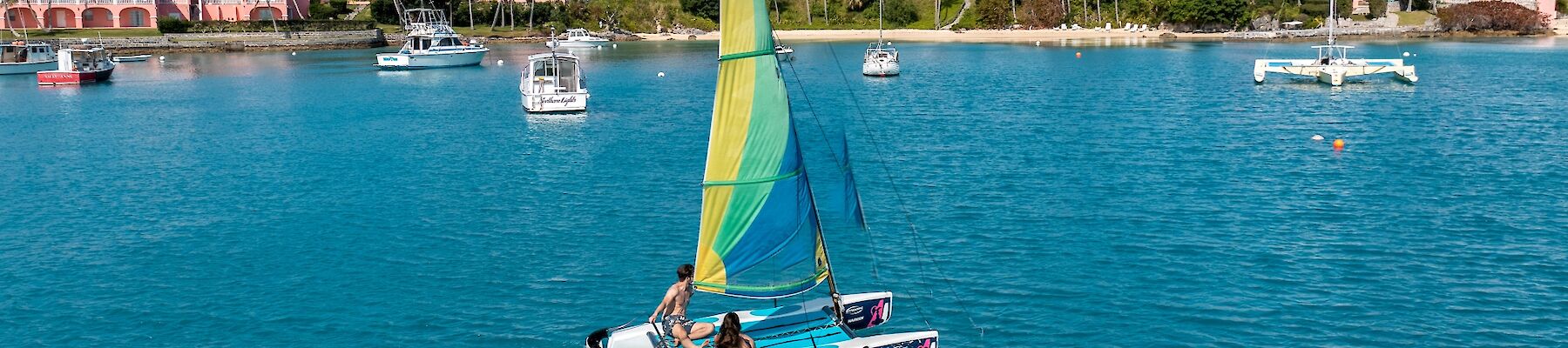 A small sailboat with a yellow and green sail glides on bright blue water near a coastal town with pink buildings and other boats in the background.