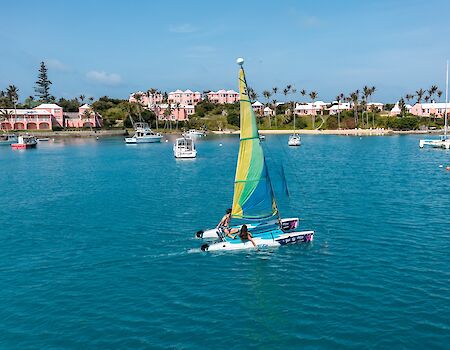A small sailboat with a yellow and green sail glides on bright blue water near a coastal town with pink buildings and other boats in the background.