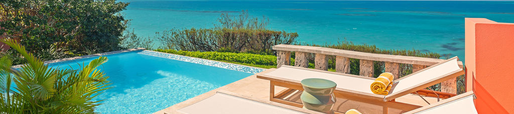 A sunny poolside with two lounge chairs, rolled towels, and a view of a turquoise sea under a clear blue sky, relaxing outdoor vibes.