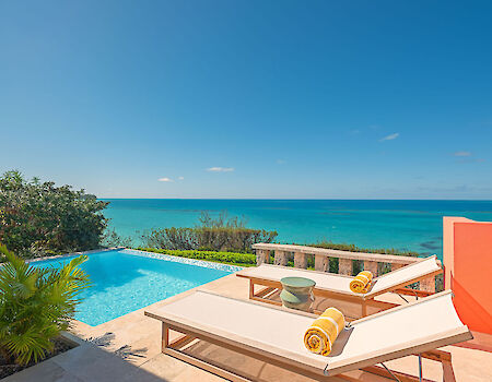 A sunny poolside with two lounge chairs, rolled towels, and a view of a turquoise sea under a clear blue sky, relaxing outdoor vibes.