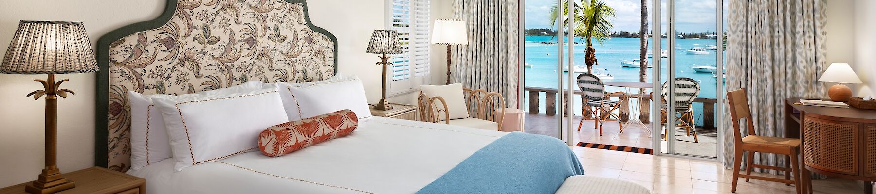 Luxurious hotel room with a decorative headboard, queen bed, blue throw, sea-view balcony, ceiling fan, and wicker furniture. End.
