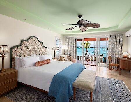 Luxurious hotel room with a decorative headboard, queen bed, blue throw, sea-view balcony, ceiling fan, and wicker furniture. End.