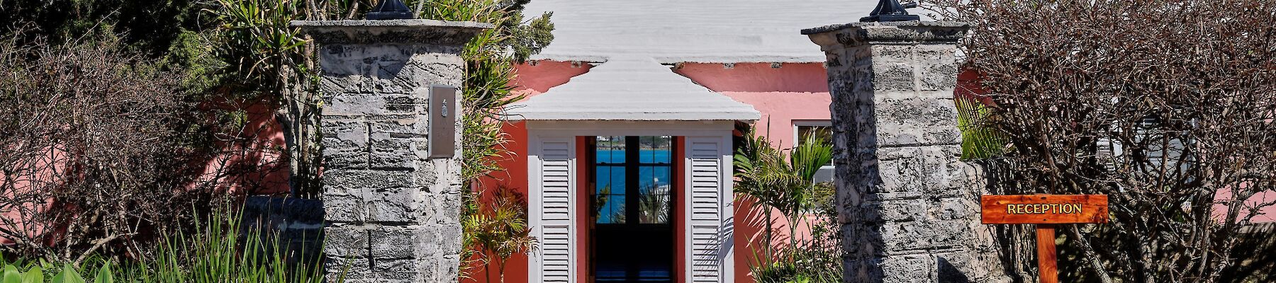 A sunny entrance to a white building with pink trim, stone pillars, tropical plants, and a welcoming path under a red awning.