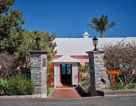 A sunny entrance to a white building with pink trim, stone pillars, tropical plants, and a welcoming path under a red awning.
