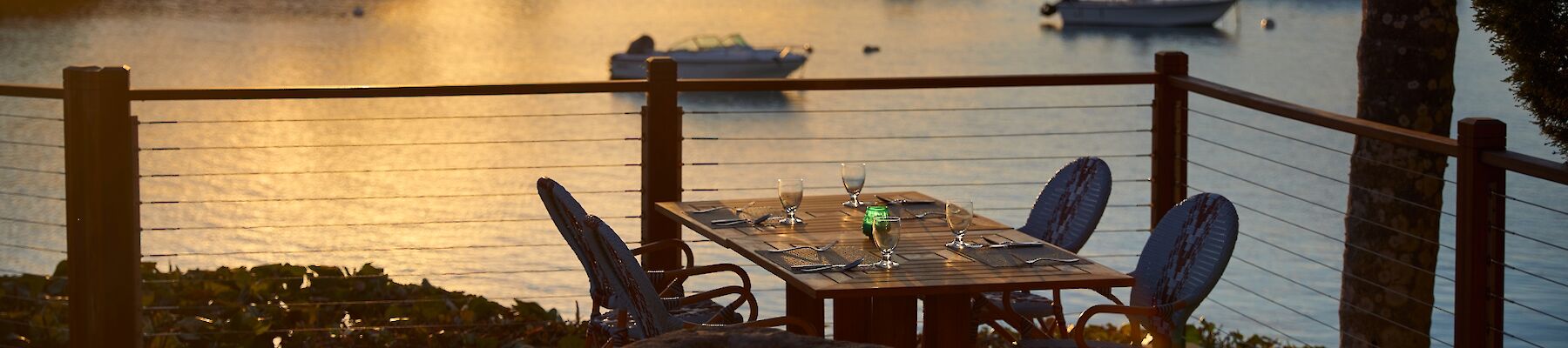 A sunset over a calm harbor with boats, palm trees, and a small patio table and chairs by the water, warm light glowing on the scene.