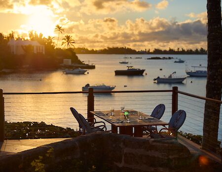 A sunset over a calm harbor with boats, palm trees, and a small patio table and chairs by the water, warm light glowing on the scene.