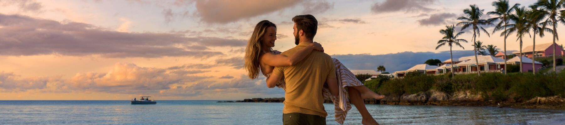 A couple on a sandy beach at sunset; the man carries a woman in his arms near calm waves, palm trees and a colorful sky in the background.