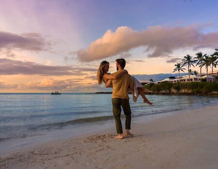 A couple on a sandy beach at sunset; the man carries a woman in his arms near calm waves, palm trees and a colorful sky in the background.