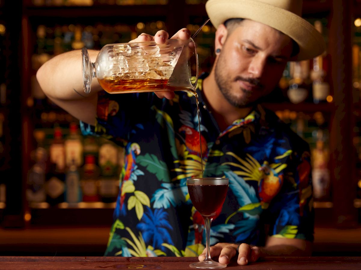 A person in a tropical shirt pours a drink from a glass container into a cocktail glass, standing in front of a bar shelf with bottles.