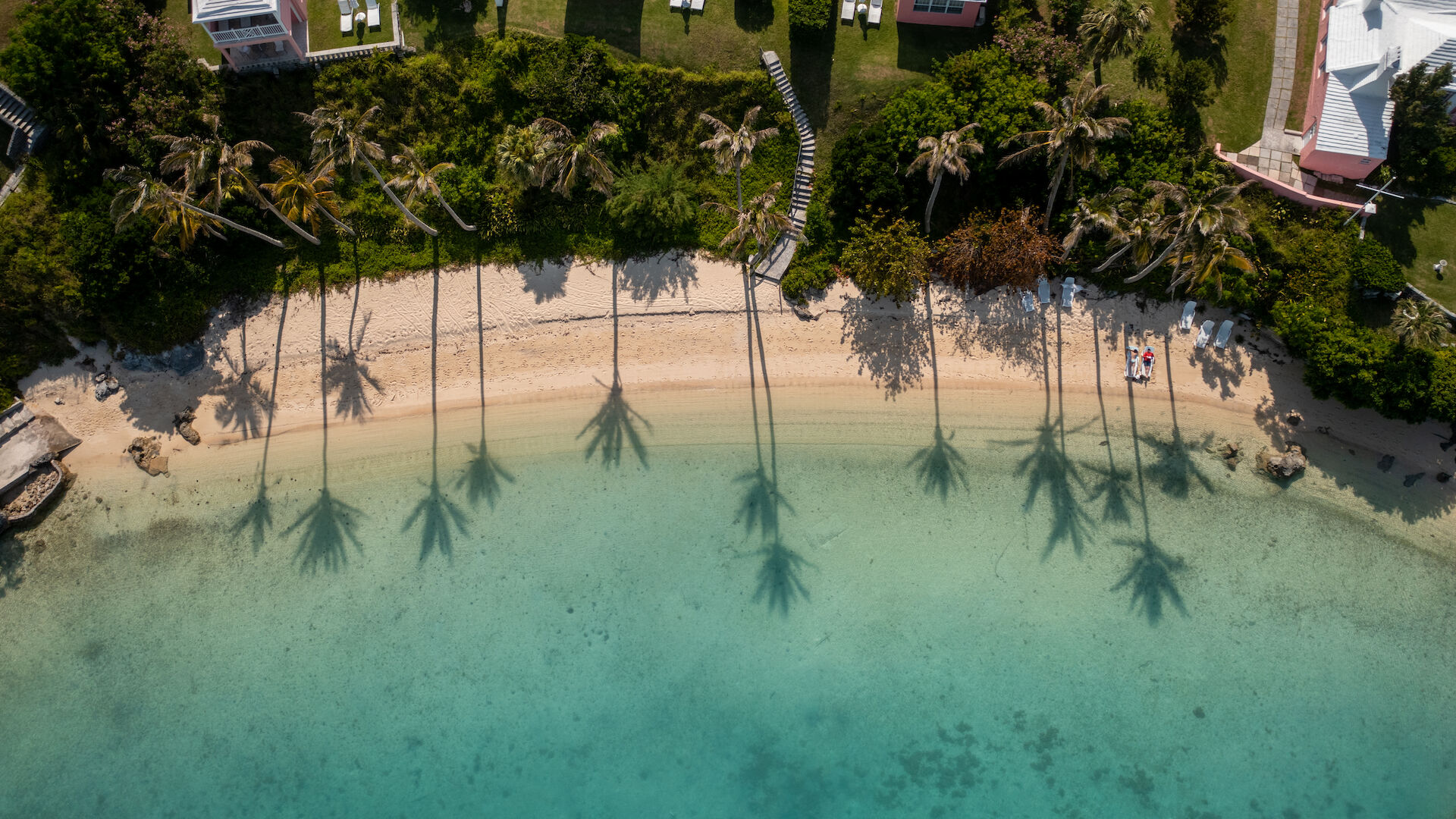 An aerial view of a beach with clear turquoise water, palm tree shadows on the sand, and houses with greenery along the shoreline, ending the sentence.