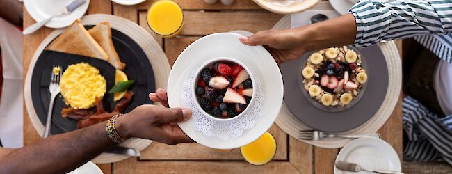 A colorful breakfast spread: waffles with eggs, berries, croissants, cereal bowls, toast, fruit, juice, and coffee on a wooden table.