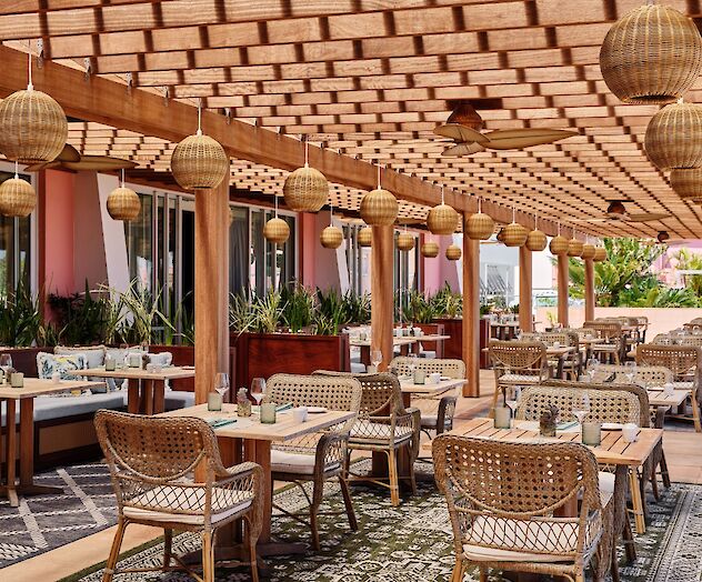 A stylish outdoor dining area with wooden beams, woven chairs, umbrellas, and lantern lighting, set with beige cushions and greenery, inviting al fresco meals.