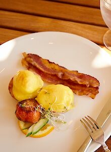 A breakfast plate with bacon, two eggs benedict, a small salad, toast, plus coffee and water on a wooden table.
