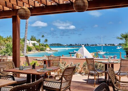 A seaside restaurant patio with wooden tables and wicker chairs overlooks a blue bay, boats anchored, and palm trees under a sunny sky.