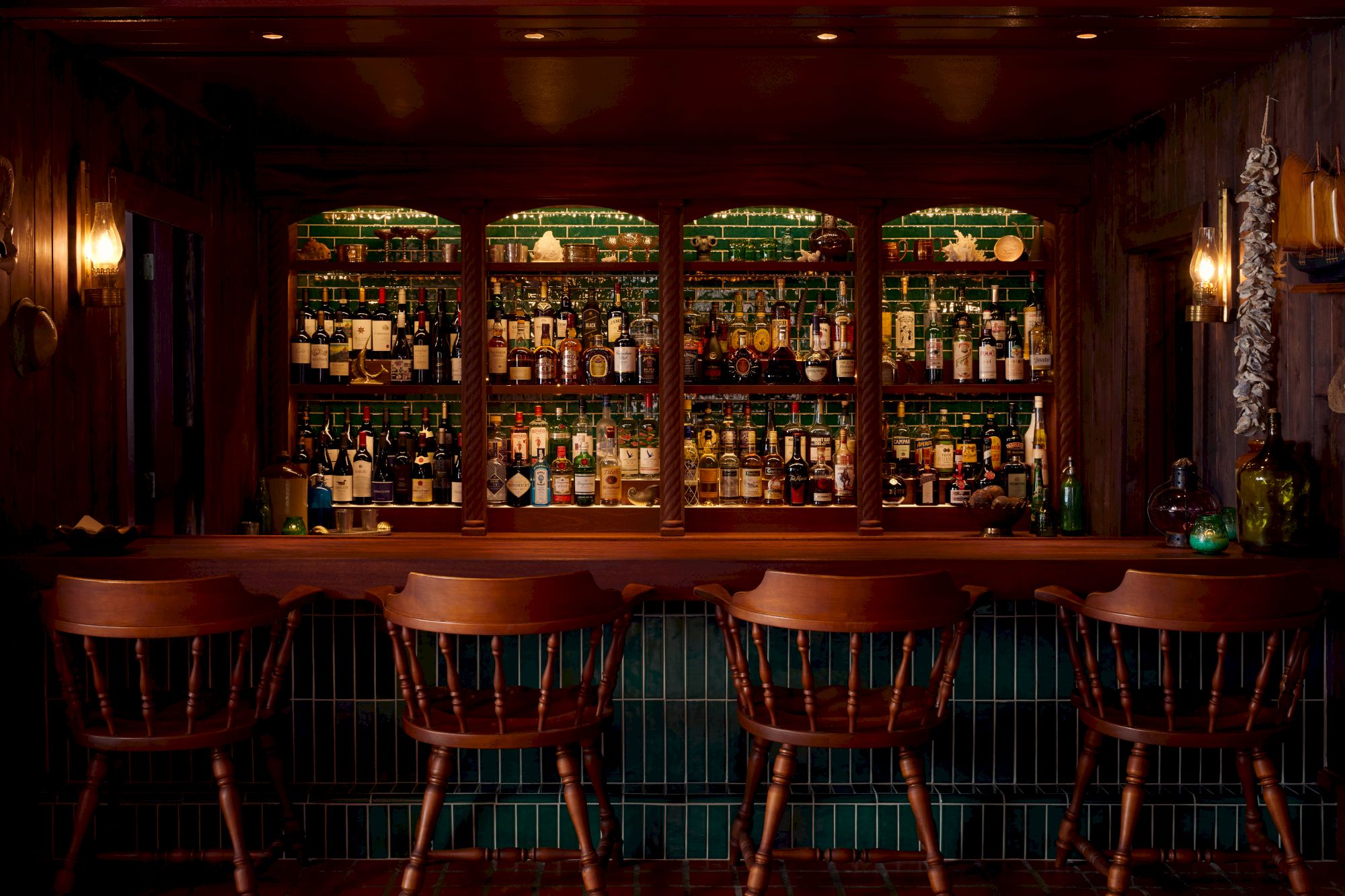 A dimly lit bar with five wooden stools, a wooden counter, and shelves filled with various bottles of alcohol in the background.