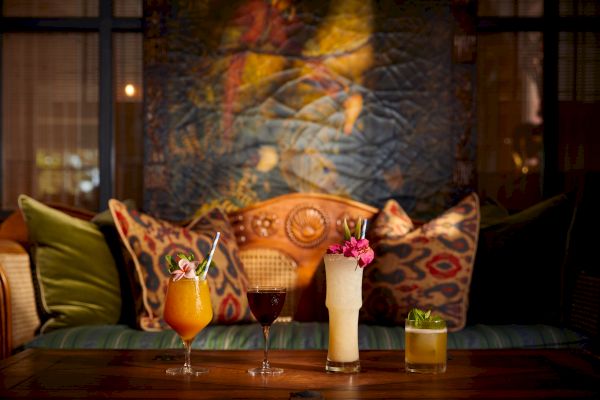 The image shows a cozy seating area with colorful patterned pillows and a wooden table featuring a variety of drinks garnished with flowers.