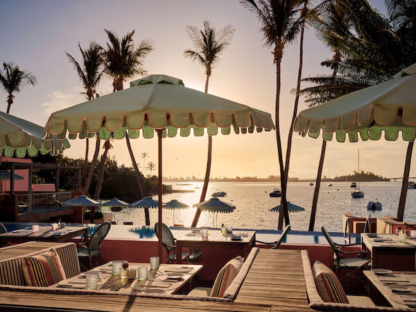 A seaside restaurant with tables, umbrellas, and palm trees overlooks a calm ocean during sunset, creating a tranquil atmosphere.